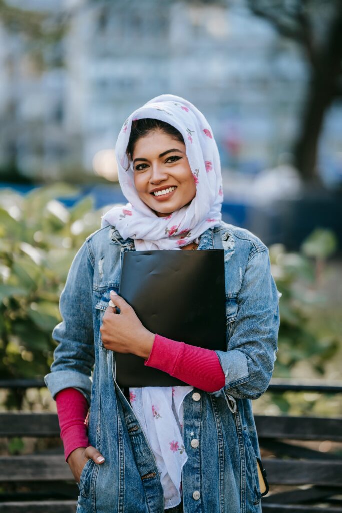 Smiling young woman in a headscarf holds a folder in a sunny park.